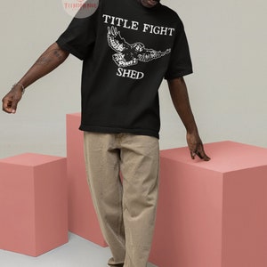 Puede incluir: Una camiseta negra con un gr&aacute;fico blanco de un b&uacute;ho volando con el texto "TITLE FIGHT SHED" impreso en la parte delantera.