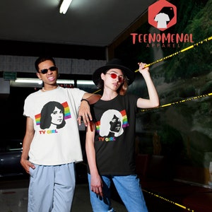 May include: Two people wearing black and white t-shirts with a silhouette of a woman's face and the text "TV GIRL" printed on them. The woman's face is outlined with a rainbow stripe. The t-shirts are being worn by two people standing in a hallway. The text "TEENOMENAL APPAREL" is visible in the background.
