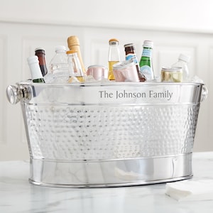 May include: A stainless steel beverage tub filled with ice and various bottles and cans. The tub has a hammered texture and the text "The Johnson Family" engraved on the side. It features two handles and is sitting on a marble surface.