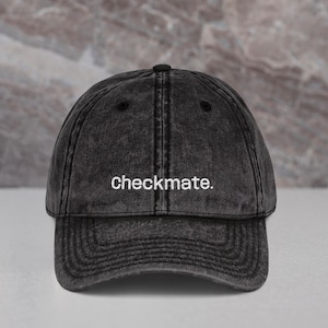 Puede incluir: Gorra de béisbol negra con la palabra "checkmate" bordada en blanco. Una pieza de ajedrez negra y una pieza de ajedrez blanca están sobre una superficie gris claro.