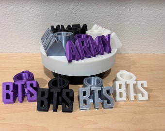 May include: A set of 3D printed words, including "BTS" in purple, black, silver, and white, and "ARMY" in purple. The words are arranged on a white rotating base.