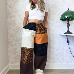 Puede incluir: Pantalones anchos con dise&ntilde;o de patchwork que incluye varios paneles, como una secci&oacute;n crema con el logo de Nike en blanco, tela marr&oacute;n, estampado de leopardo y tela negra con lunares blancos. Los pantalones tienen cintura el&aacute;stica y corte holgado.