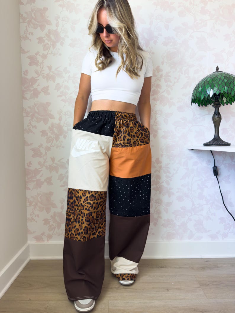 Puede incluir: Pantalones anchos con dise&ntilde;o de patchwork que incluye varios paneles, como una secci&oacute;n crema con el logo de Nike en blanco, tela marr&oacute;n, estampado de leopardo y tela negra con lunares blancos. Los pantalones tienen cintura el&aacute;stica y corte holgado.
