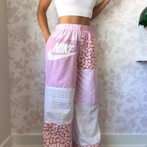 Puede incluir: Pantalones de patchwork de talle alto con tejido rosa claro y un logotipo de Nike blanco en el muslo derecho, alternando con secciones de tela de encaje blanco y un estampado floral rosa con peque&ntilde;as margaritas blancas en las piernas.