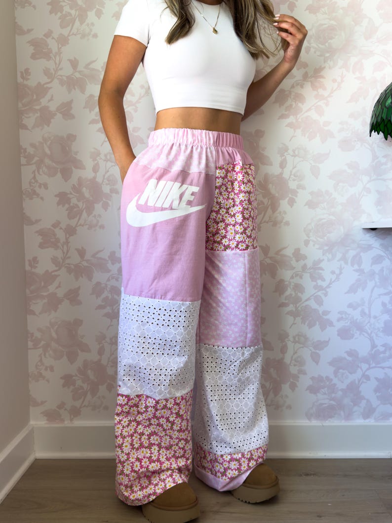 Puede incluir: Pantalones de patchwork de talle alto con tejido rosa claro y un logotipo de Nike blanco en el muslo derecho, alternando con secciones de tela de encaje blanco y un estampado floral rosa con peque&ntilde;as margaritas blancas en las piernas.