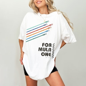 Könnte beinhalten: Weißes Oversize-T-Shirt mit einem Aufdruck von bunten Rennstreifen und Autos. Der Text "FOR MULA ONE est. 1950" ist auf das Shirt gedruckt.