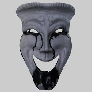 May include: A gray theatrical mask with a smiling mouth and open eye sockets. Black paint drips from the eyes and mouth, creating a visual contrast. The mask has a textured surface and a decorative top.