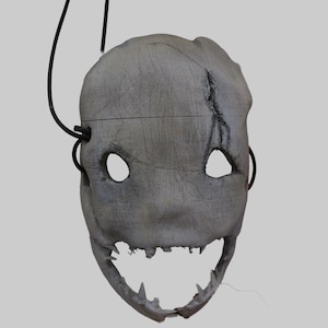 May include: A grey, textured mask featuring two oval eye openings, a jagged, toothy mouth opening, and a prominent crack detail on the upper right side. A thin, black, curved wire extends from the left side of the mask, arching upwards.