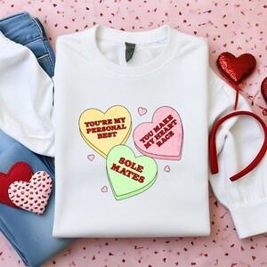 May include: White sweatshirt with a graphic of three candy hearts. The hearts say "You're my personal best", "You make my heart race", and "Sole Mates".