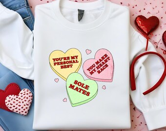 May include: White sweatshirt with a graphic of three candy hearts. The hearts say "You're my personal best", "You make my heart race", and "Sole Mates".