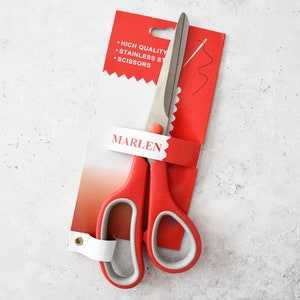 Könnte beinhalten: Rote Stoffschere mit einer gezahnten Klinge und einem weißen Kunststoffgriff. Die Schere ist auf einer roten Karte verpackt mit dem Text "HIGH QUALITY STAINLESS STEEL SCISSORS" und "MARLEN".