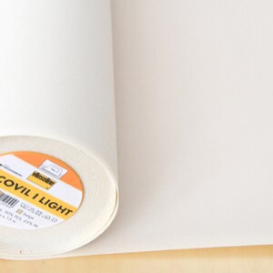 Puede incluir: Un rollo de entretela fusible Decovil I Light blanco. La etiqueta es amarilla con texto negro y un código de barras. El texto de la etiqueta dice "Decovil I Light" y "25 cm x 25 m".