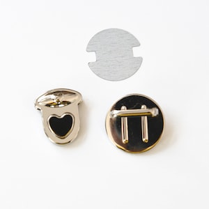 Op de afbeelding: Drie zilveren metalen sluitingen om kleding of accessoires aan te bevestigen. Een sluiting heeft een hartvormig ontwerp met een zwart midden. Een andere sluiting heeft een eenvoudig, rond ontwerp met twee parallelle balken. De derde sluiting is een rond, plat stuk metaal met een uitsparing in het midden.
