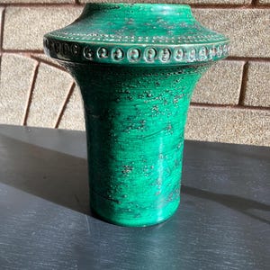 May include: A green ceramic vase with a textured surface. The vase has a wide, decorative rim with a series of small, circular designs. The body of the vase is cylindrical, with a slightly flared top. The vase is approximately 10 inches tall.