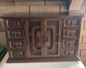 May include: A dark brown wooden jewelry armoire featuring two central doors with geometric paneling and eight small drawers, four on each side. Each drawer and door is equipped with a small, vertical, dark metal pull handle for access.