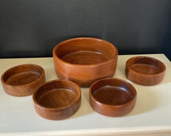 Peut inclure: Un ensemble de cinq bols en bois de différentes tailles. Les bols sont de couleur brun chaud, avec un grain de bois visible. Le plus grand bol est au centre, entouré de quatre bols plus petits. Les bols semblent faits à la main.