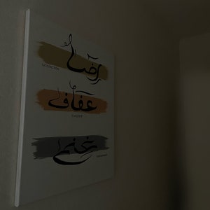 May include: A white canvas art piece with Arabic calligraphy and English translations. The artwork features three horizontal brushstrokes in gold, orange, and grey, each with a word in Arabic script and its English translation: "Satisfaction", "Chastete", and "Contentment".