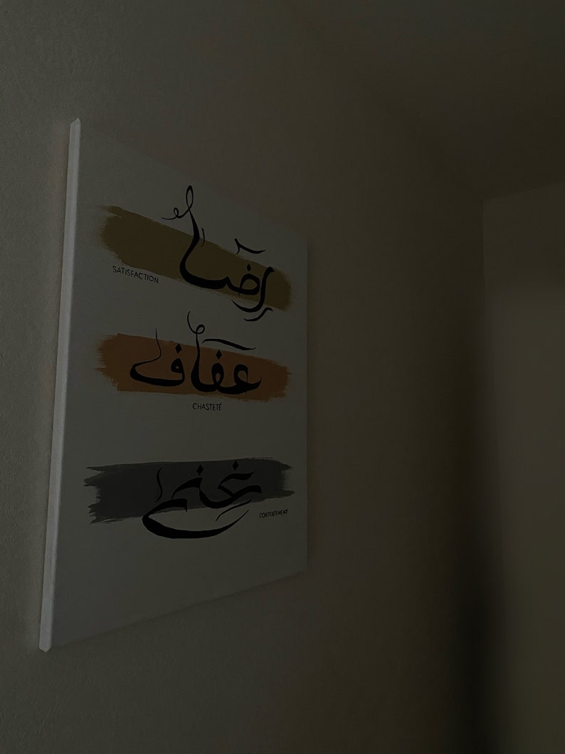 May include: A white canvas art piece with Arabic calligraphy and English translations. The artwork features three horizontal brushstrokes in gold, orange, and grey, each with a word in Arabic script and its English translation: "Satisfaction", "Chastete", and "Contentment".