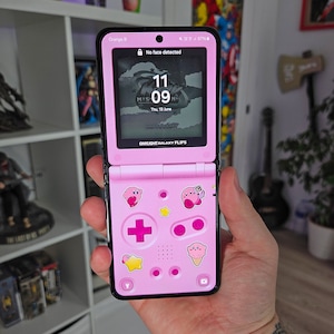 May include: A pink and black flip phone with a pink Game Boy Advance SP-style case. The phone has a black screen with the time displayed as 11:09. The case has a pink and white star, a pink plus sign, and a pink and white ice cream cone with a pink and white Kirby character on it.