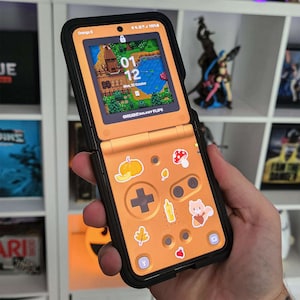 May include: An orange and black smartphone case with a retro video game controller design. The phone screen displays a landscape scene with a clock showing 01:12 and the date. Decorative stickers include a pumpkin, mushroom, and leaf.