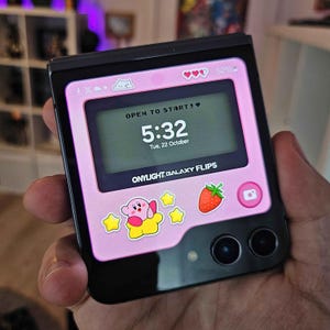 以下が含まれることがあります： ピンクとブラックのGalaxy Flip5電話は、ピンクとホワイトのカービィキャラクター、イチゴ、星が描かれたカスタムケースが付いています。電話の画面には、時間「5:32」と日付「Tue, 22 October」が表示されています。画面には、「OPEN TO START!❤」と「ONYLIGHT. GALAXY FLIP5」のテキストも表示されています。