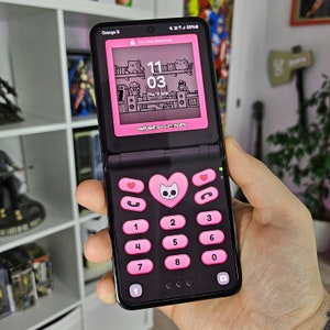 以下が含まれることがあります： ピンクのハート型のボタンと、11:03 の時間を表示するデジタル時計が付いた黒いフリップフォン。電話には、猫のピクセル化された画像が描かれた白黒の画面があります。画面には「ONYLIGHT GALAXY FLIPS」というテキストが表示されています。