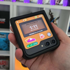 Può includere: Una custodia per telefono nera con un proteggi schermo dorato e arancione. Il proteggi schermo ha un orologio, una data e il testo "OPEN TO START!" e "ONYLIGHT.GALAXY FLIPS".