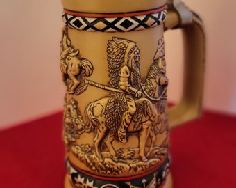 May include: Tan ceramic beer stein with a detailed relief of Native American figures on horseback. The lid is adorned with a turquoise-colored design and a metal hinge. The handle is tan, and the base features a black, red, and green pattern.