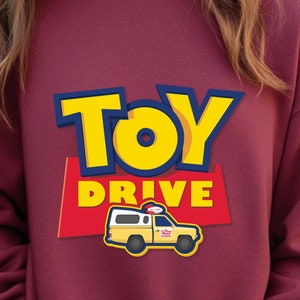 May include: A maroon sweatshirt with a yellow and blue graphic of the words "TOY DRIVE" and a cartoon yellow van with a red roof.