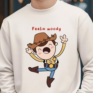 Puede incluir: Una sudadera de color crema con una ilustraci&oacute;n de dibujos animados de Woody de Toy Story. El texto "Feelin' Woody" est&aacute; impreso en rojo sobre la ilustraci&oacute;n.
