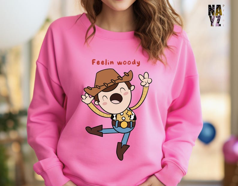 Puede incluir: Sudadera rosa con una ilustraci&oacute;n de dibujos animados de Woody de Toy Story. La ilustraci&oacute;n muestra a Woody con una gran sonrisa y los brazos en alto. El texto "Feelin' Woody" est&aacute; impreso sobre la ilustraci&oacute;n.