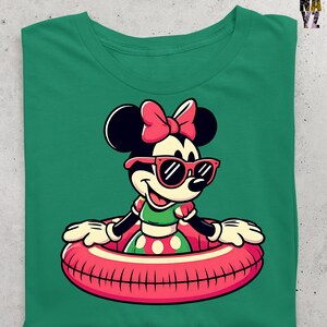 May include: Green t-shirt with a cartoon character wearing sunglasses and a pink bow. The character is in a red and pink inner tube. The character is wearing a green top and a polka dot skirt. The shirt has a crew neck.