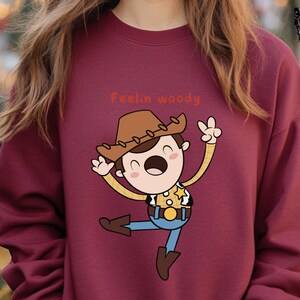 Puede incluir: Una sudadera color burdeos con una ilustraci&oacute;n de dibujos animados de Woody de Toy Story. La ilustraci&oacute;n muestra a Woody con su caracter&iacute;stico sombrero de vaquero, chaleco y una expresi&oacute;n alegre. El texto "Feelin' Woody" est&aacute; impreso sobre la ilustraci&oacute;n.