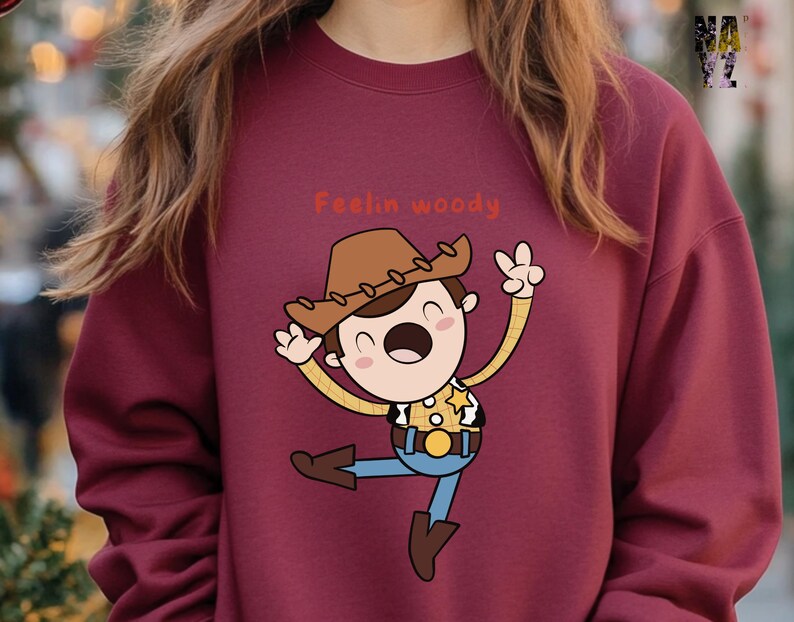 Puede incluir: Una sudadera color burdeos con una ilustraci&oacute;n de dibujos animados de Woody de Toy Story. La ilustraci&oacute;n muestra a Woody con su caracter&iacute;stico sombrero de vaquero, chaleco y una expresi&oacute;n alegre. El texto "Feelin' Woody" est&aacute; impreso sobre la ilustraci&oacute;n.