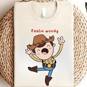 Puede incluir: Una camiseta blanca con una ilustraci&oacute;n de dibujos animados de Woody de Toy Story. La ilustraci&oacute;n muestra a Woody con su caracter&iacute;stico sombrero de vaquero, chaleco y una expresi&oacute;n alegre. El texto "Feelin' Woody" est&aacute; impreso sobre la ilustraci&oacute;n.
