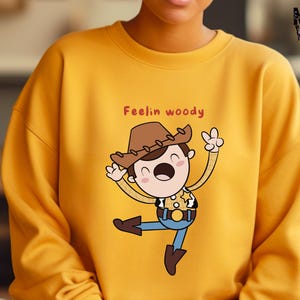 Puede incluir: Sudadera amarilla con una ilustraci&oacute;n de dibujos animados de Woody de Toy Story. El texto "Feelin' Woody" est&aacute; impreso en la sudadera.