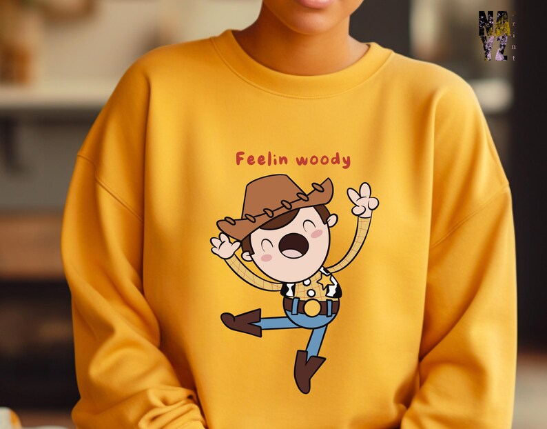Puede incluir: Sudadera amarilla con una ilustraci&oacute;n de dibujos animados de Woody de Toy Story. El texto "Feelin' Woody" est&aacute; impreso en la sudadera.