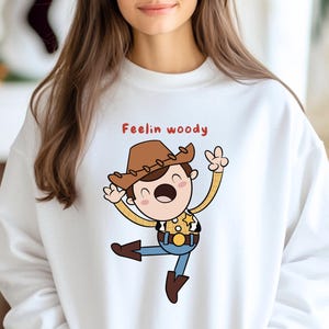 Puede incluir: Sudadera blanca con una ilustraci&oacute;n de dibujos animados de Woody de Toy Story. El texto "Feelin Woody" est&aacute; impreso en rojo sobre la ilustraci&oacute;n.