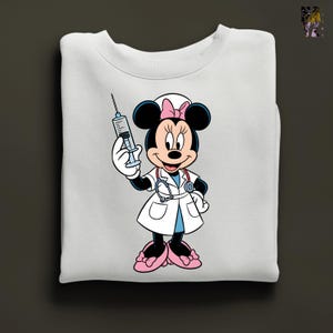 Peut inclure: Sweat-shirt blanc avec Minnie Mouse en infirmi&egrave;re, tenant une seringue. Minnie Mouse porte une blouse blanche, des chaussures roses et un n&oelig;ud rose. Le sweat-shirt est pli&eacute;.