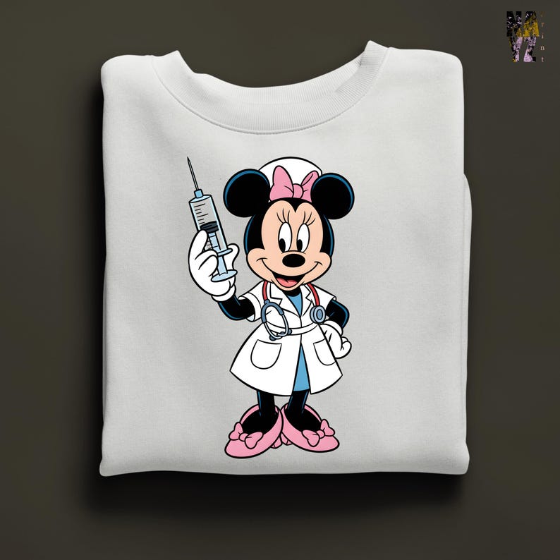 Peut inclure: Sweat-shirt blanc avec Minnie Mouse en infirmi&egrave;re, tenant une seringue. Minnie Mouse porte une blouse blanche, des chaussures roses et un n&oelig;ud rose. Le sweat-shirt est pli&eacute;.