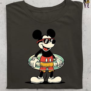 May include: Dark gray t-shirt featuring a cartoon Mickey Mouse character wearing sunglasses and swim trunks, holding a striped inflatable ring. The character is smiling and ready for summer fun. The shirt has a classic, vintage design.