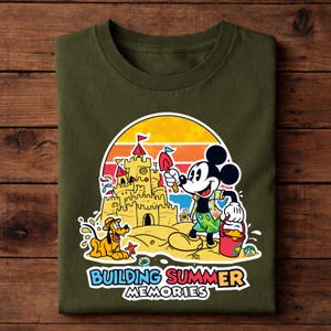 May include: Olive green t-shirt featuring a colorful graphic of Mickey Mouse building a sandcastle with Pluto. The design includes a sunset backdrop and the text "Building Summer Memories."
