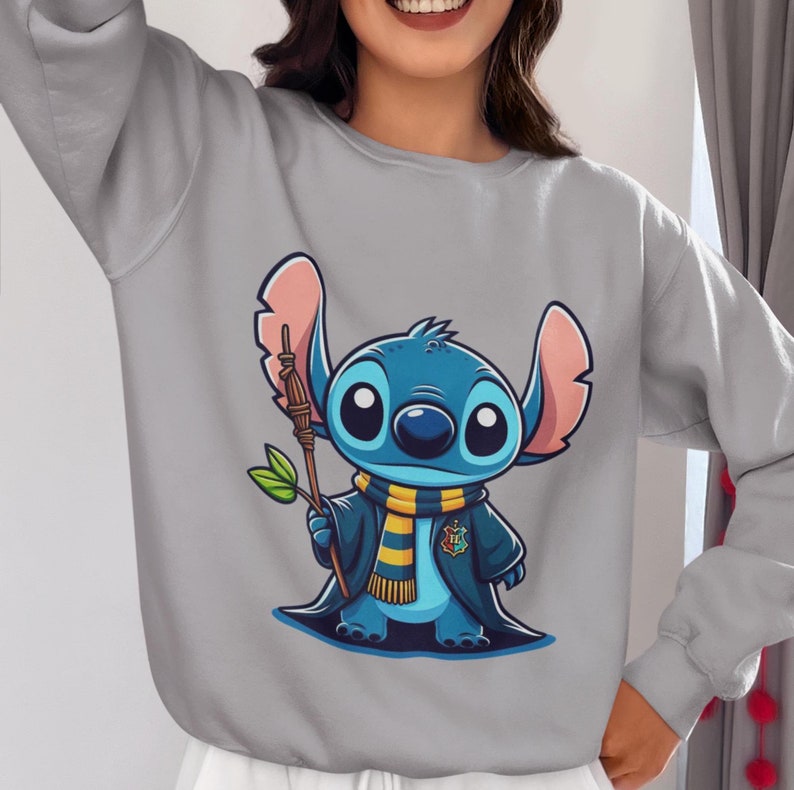 May include: Gray sweatshirt with a blue Stitch character wearing a yellow and blue scarf and holding a brown stick. Stitch is wearing a blue robe with a yellow and red crest.