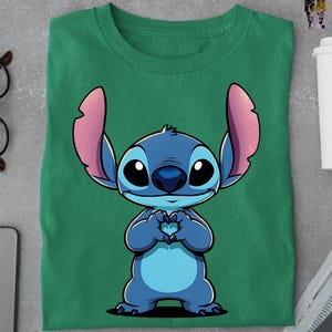 May include: Green t-shirt featuring a cartoon illustration of Stitch from Lilo & Stitch. The blue character is making a heart shape with its hands. The shirt is folded and has a crew neck.