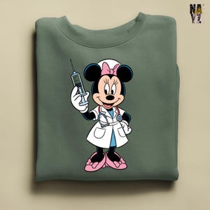 Peut inclure: Sweat-shirt vert sauge avec Minnie Mouse en infirmi&egrave;re, tenant une seringue. Minnie porte une blouse blanche, des chaussures roses et un bonnet d'infirmi&egrave;re avec un n&oelig;ud rose. Le sweat-shirt a un col rond.