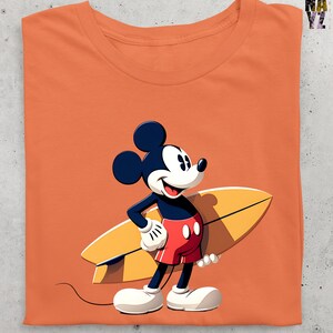 Op de afbeelding: Oranje t-shirt met een cartoonafbeelding van Mickey Mouse met een geel surfboard. Mickey draagt een rode korte broek, witte handschoenen en schoenen. Het shirt heeft een ronde hals en is opgevouwen.