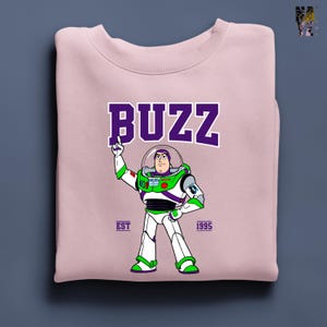 May include: A light pink sweatshirt featuring a graphic of Buzz Lightyear from Toy Story. The word "BUZZ" is printed in purple block letters above the character. The sweatshirt also includes the text "EST 1995".