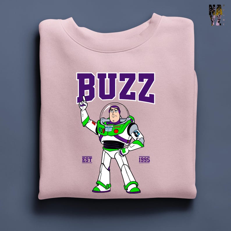 May include: A light pink sweatshirt featuring a graphic of Buzz Lightyear from Toy Story. The word "BUZZ" is printed in purple block letters above the character. The sweatshirt also includes the text "EST 1995".
