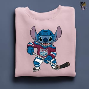Puede incluir: Sudadera rosa claro con un personaje de dibujos animados Stitch con uniforme de hockey. Stitch lleva un casco azul y sostiene un palo de hockey. El uniforme es burdeos, azul y blanco, con un logotipo del equipo en el pecho.