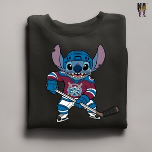 Puede incluir: Sudadera negra con un personaje de dibujos animados con uniforme de hockey. El personaje azul con orejas grandes lleva un casco. La camiseta de hockey es granate, azul y blanca, con un logotipo del equipo. Sostiene un palo de hockey.